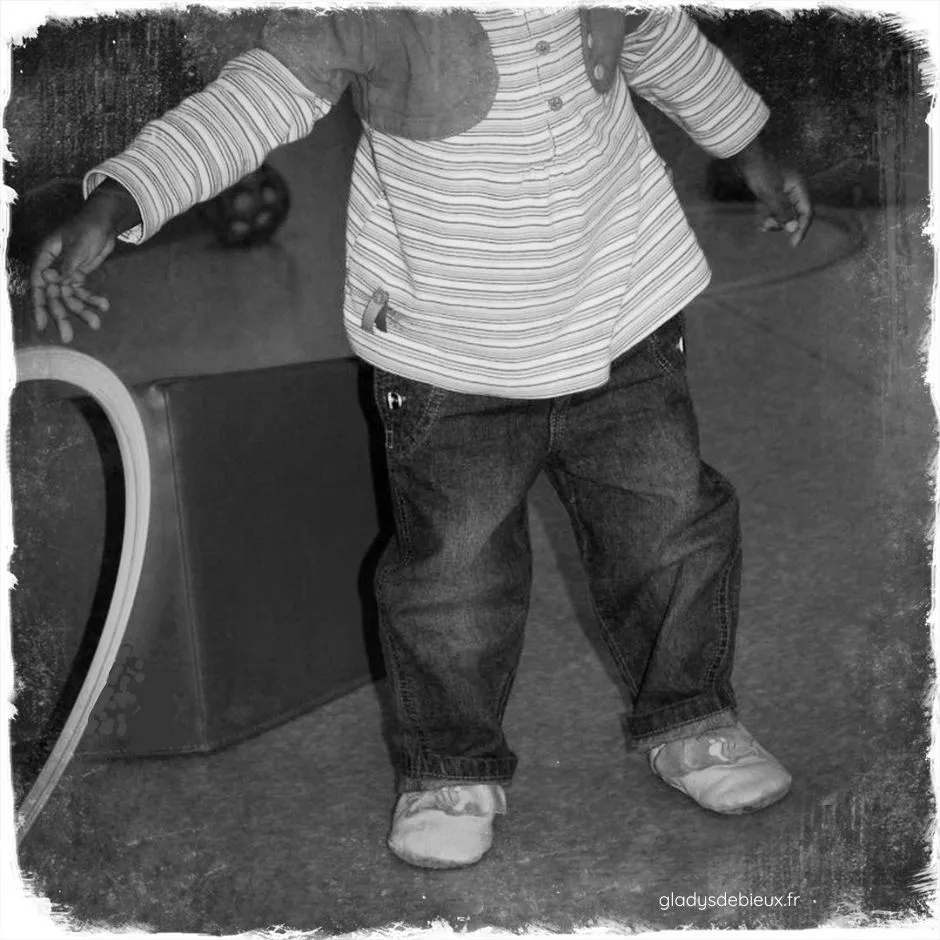Photo noir et blanc d'un enfant debout jouant avec un cerceau. Nous ne distinguons pas son visage, seulement le reste de son corps. Le but de l'image est de montrer les chaussons souples qu'elle porte aux pieds.